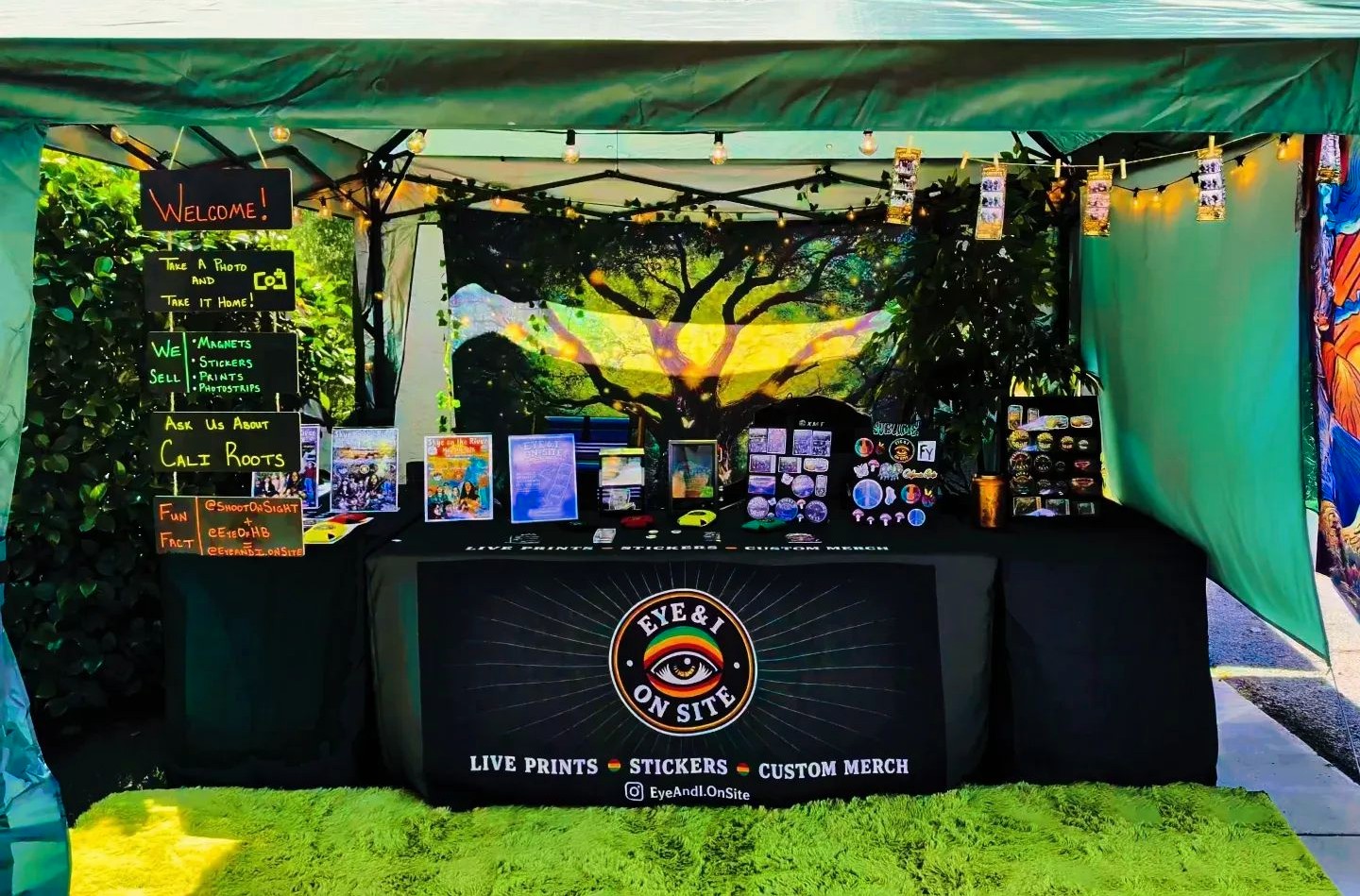 The Eye and I On-Site booth at dusk: branded table with live prints, stickers, and custom merch under a lit tent