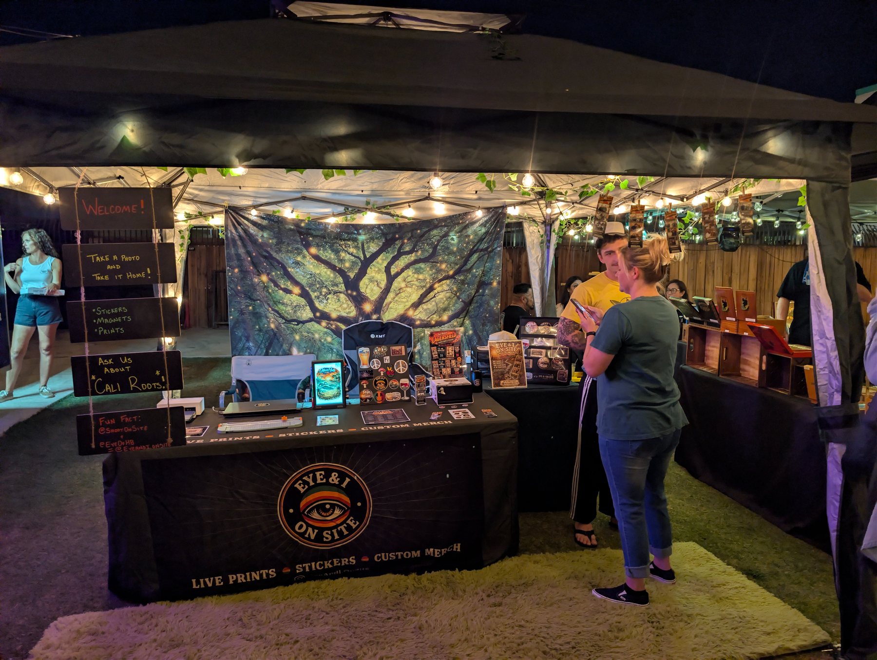 The Eye & I On-Site booth at Lagersfield with string lights, a tapestry backdrop, sticker displays, and guests browsing merch